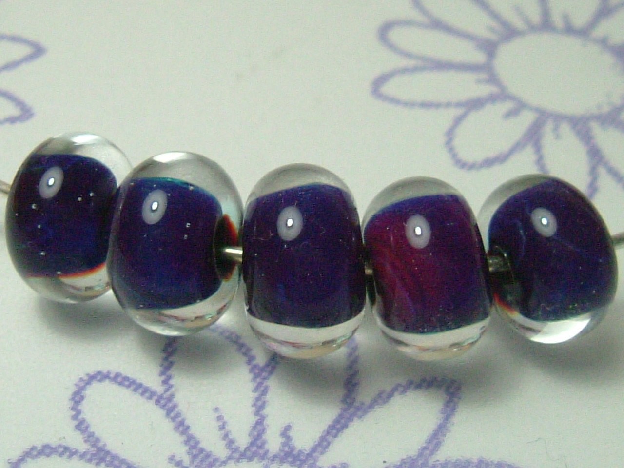 A set of 5 beads, made using reactive glass which has given shades of mainly purples and blues. Encased with a thick layer of crystal clear glass.
